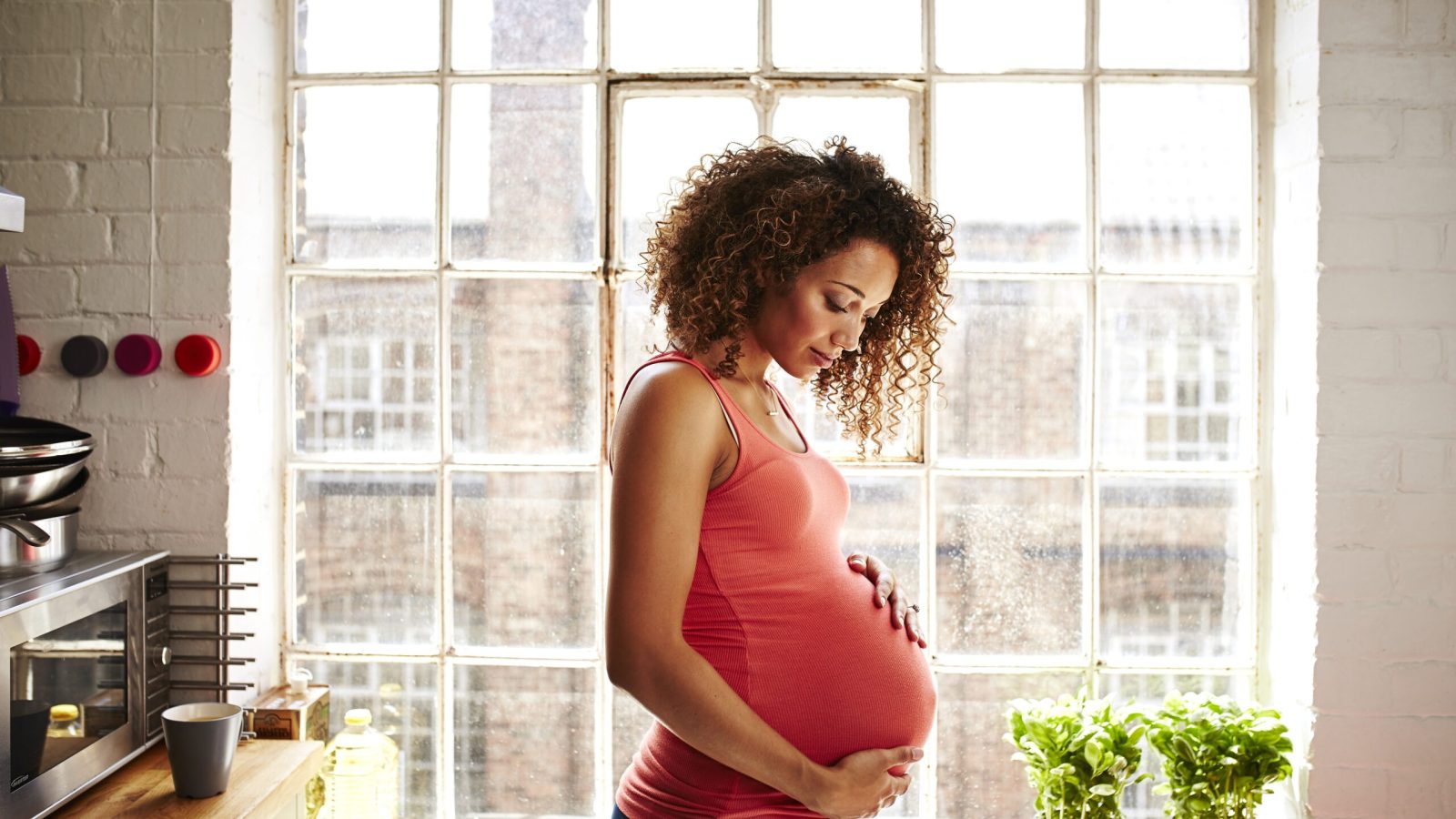 A pregnant woman holds her bump thoughtfully in her modern kitchen window on a sunny day