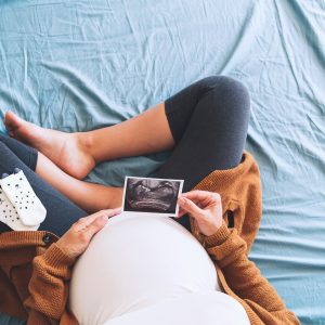 Pregnant woman holding ultrasound image. Concept of pregnancy, health care, gynecology, medicine. Young mother waiting of the baby. Close-up, copy space, indoors.
